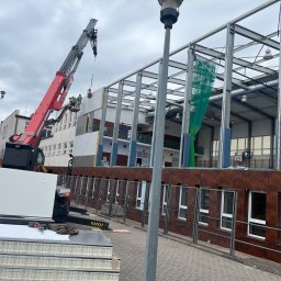 Mateusz Laskowski - Montaż paneli ściennych na stalowej konstrukcji hali sportowej z użyciem dźwigu. Widoczni pracownicy w kaskach. Na pierwszym planie stos paneli i narzędzia.