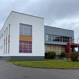 Mateusz Laskowski - Dwukondygnacyjny budynek z białymi i szarymi panelami, z szyldem 'Cafe Muza' na oknie. Widok z zewnątrz, z chodnikiem i zielenią.