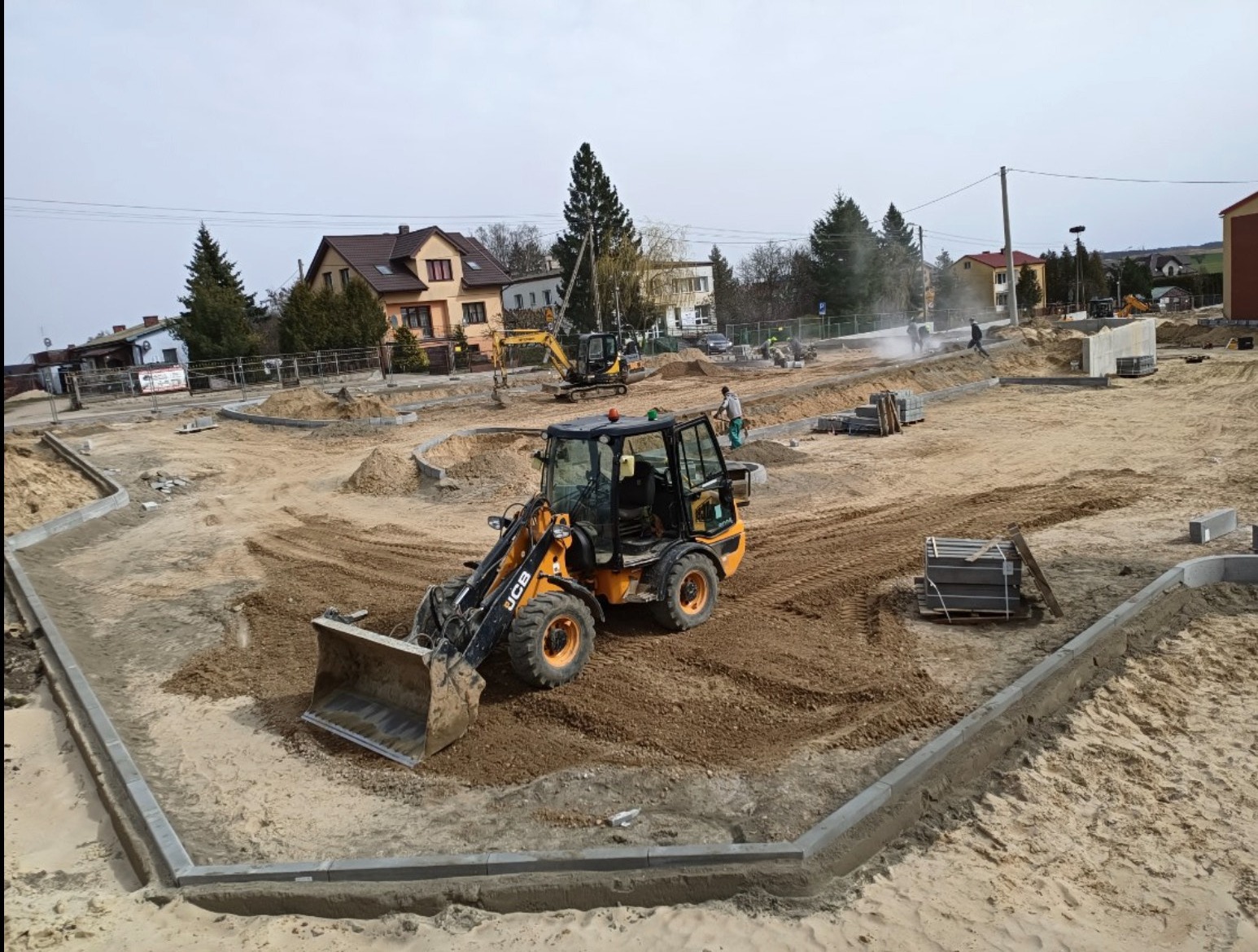 Żółta koparko-ładowarka JCB na placu budowy z ułożonymi krawężnikami i piaskiem. W tle inne maszyny i pracownicy. Etap przygotowania terenu pod inwestycję.