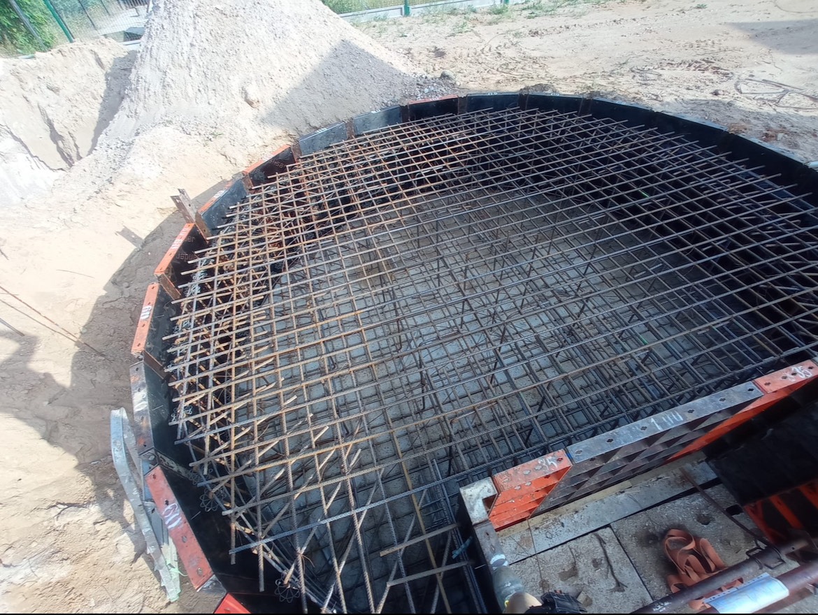 Zbrojenie okrągłego fundamentu w szalunku, widok z góry. Siatka z prętów stalowych, piasek w tle. Etap budowy, przygotowanie do zalewania betonem.