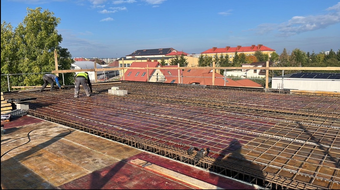 Zbrojenie stropu na budowie z widocznymi pracownikami i elementami konstrukcyjnymi. W tle budynki mieszkalne i błękitne niebo.
