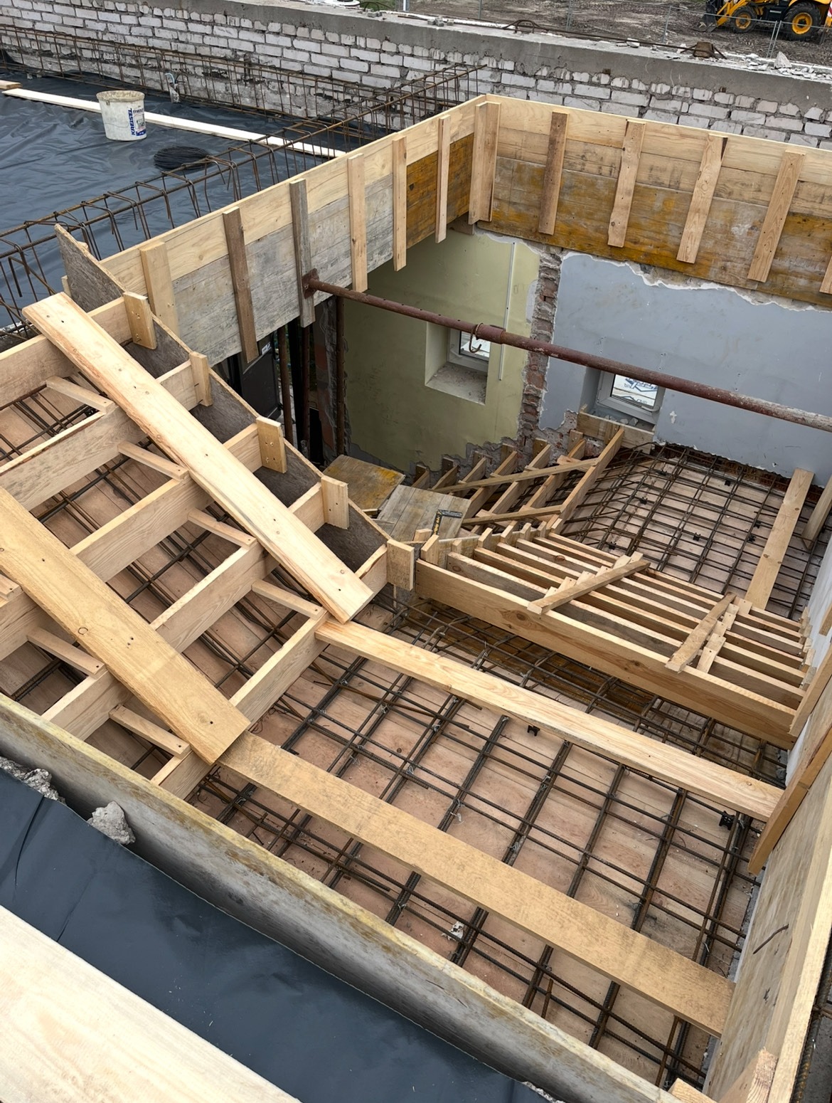 Zbrojenie i szalunek schodów w trakcie budowy. Widoczne drewniane elementy konstrukcyjne i siatka zbrojeniowa. W tle surowy mur i okno.