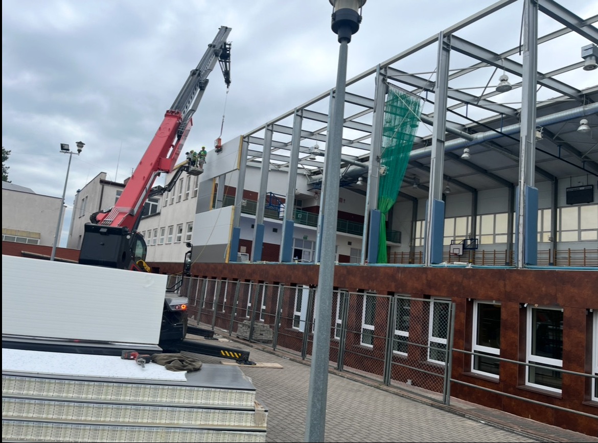 Montaż paneli ściennych na konstrukcji stalowej hali sportowej z użyciem dźwigu. Widoczni pracownicy w koszu dźwigu. Częściowo widoczna elewacja budynku.
