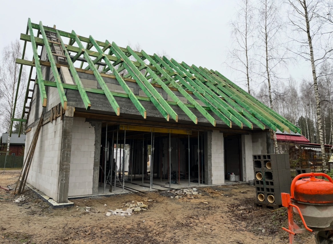 Budowa domu jednorodzinnego w trakcie realizacji: ściany z bloczków, zielona więźba dachowa, widoczna betoniarka i pustaki wentylacyjne. Surowy stan budynku.