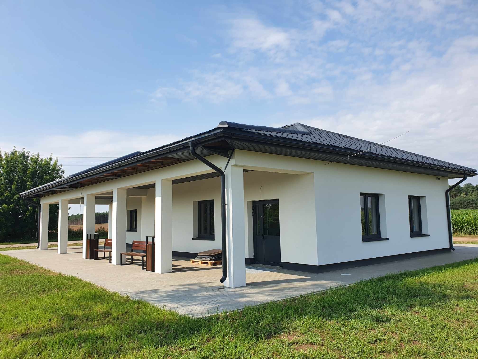Biały budynek z kolumnami i ciemną dachówką, widok z zewnątrz, błękitne niebo w tle. Na podłożu widoczne płyty chodnikowe i trawa.