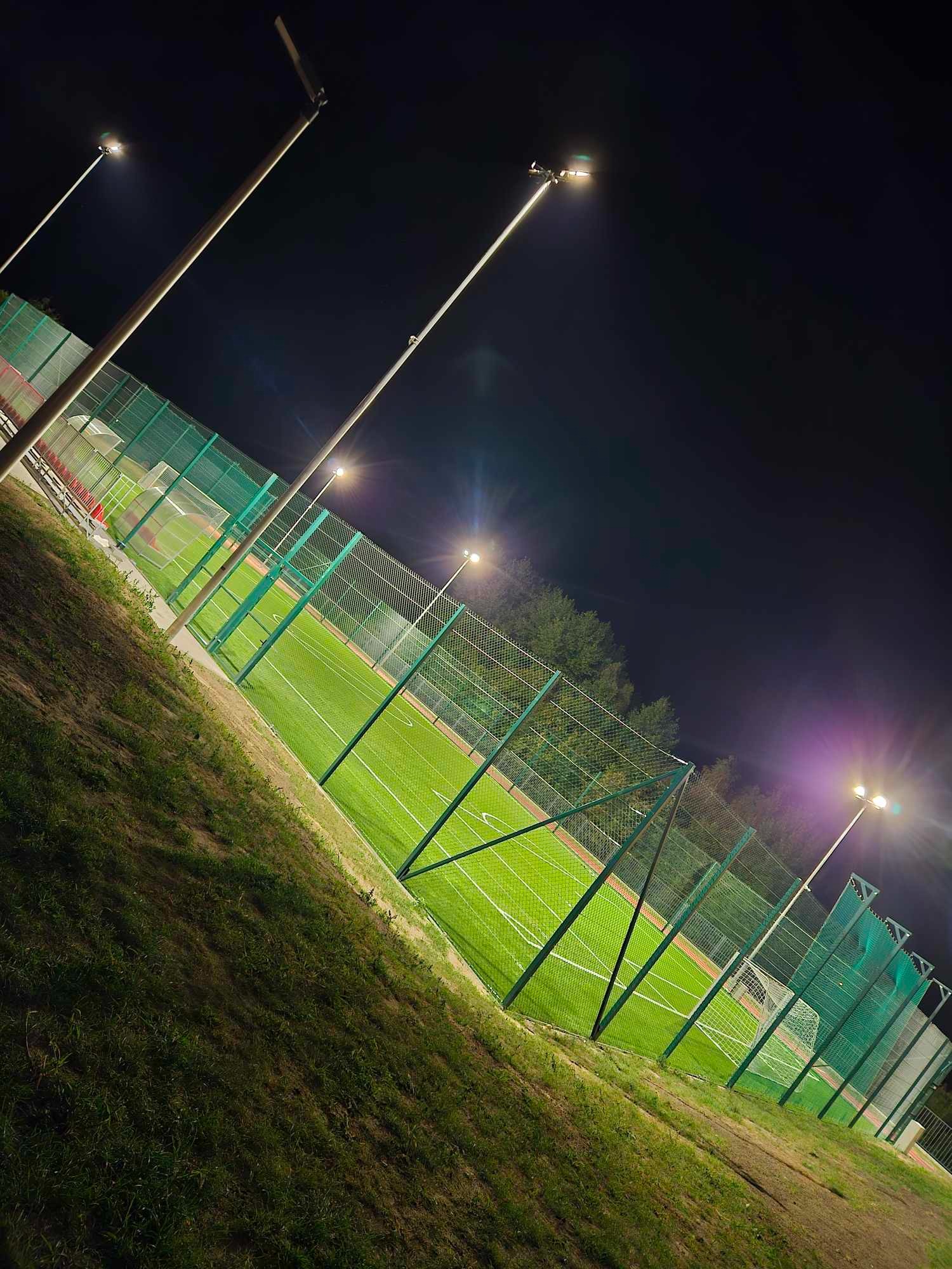 Oświetlone boisko sportowe z zieloną trawą i siatką ochronną, widoczne w nocy pod kątem. Lampy oświetlają teren, a w tle widać drzewa.