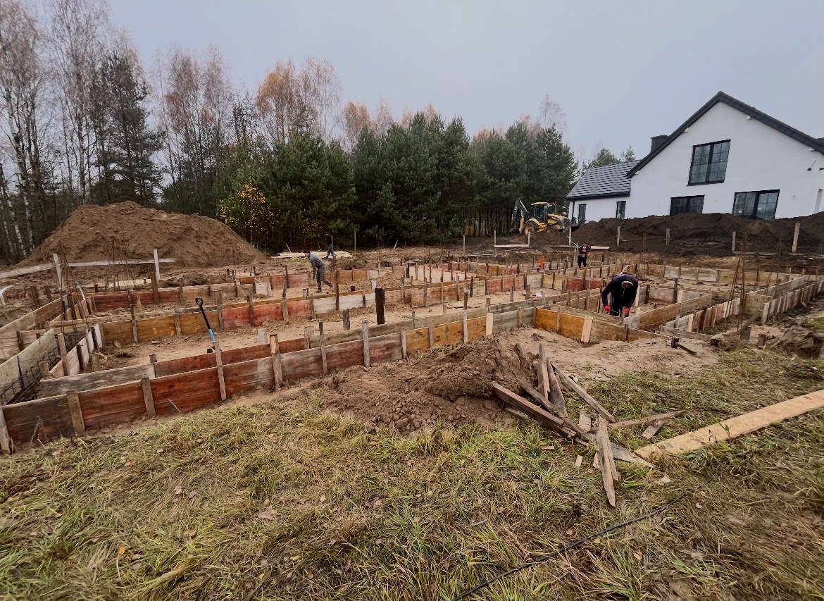 Budowa fundamentów domu jednorodzinnego: szalunki z desek, wykop, pracownicy w trakcie robót ziemnych, koparka w tle, dom w stanie surowym otwartym.