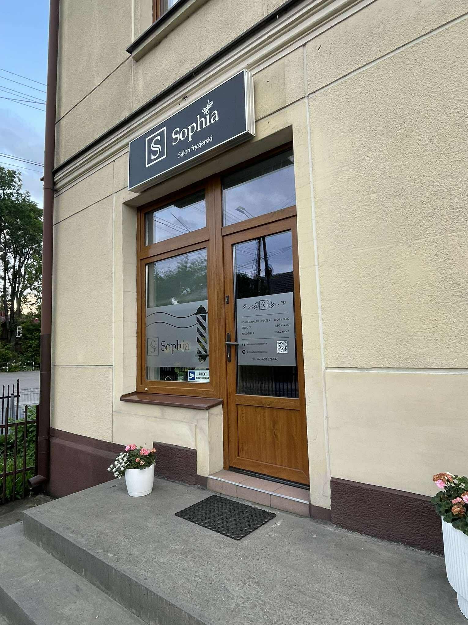 Fasada salonu fryzjerskiego Sophia z szyldem i okleiną okienną z logo. Wejście zdobią kwiaty w donicach. Ujęcie z dołu, podkreślające detale elewacji.