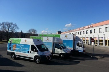 Trzy busy transportowe Home Service zaparkowane przed budynkiem. Pierwszy bus z grafiką przedstawiającą pakowanie. Błękitne niebo, słoneczny dzień.