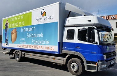 Ciężarówka Home-Service z reklamą transportu, załadunku i pakowania, zaparkowana przed budynkiem. Widoczny numer telefonu i adres strony internetowej.