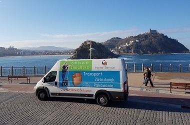 Biały bus z grafiką transportową na tle morza i górzystego krajobrazu. Widoczny napis 'Transport Załadunek Pakowanie' i dane kontaktowe firmy.