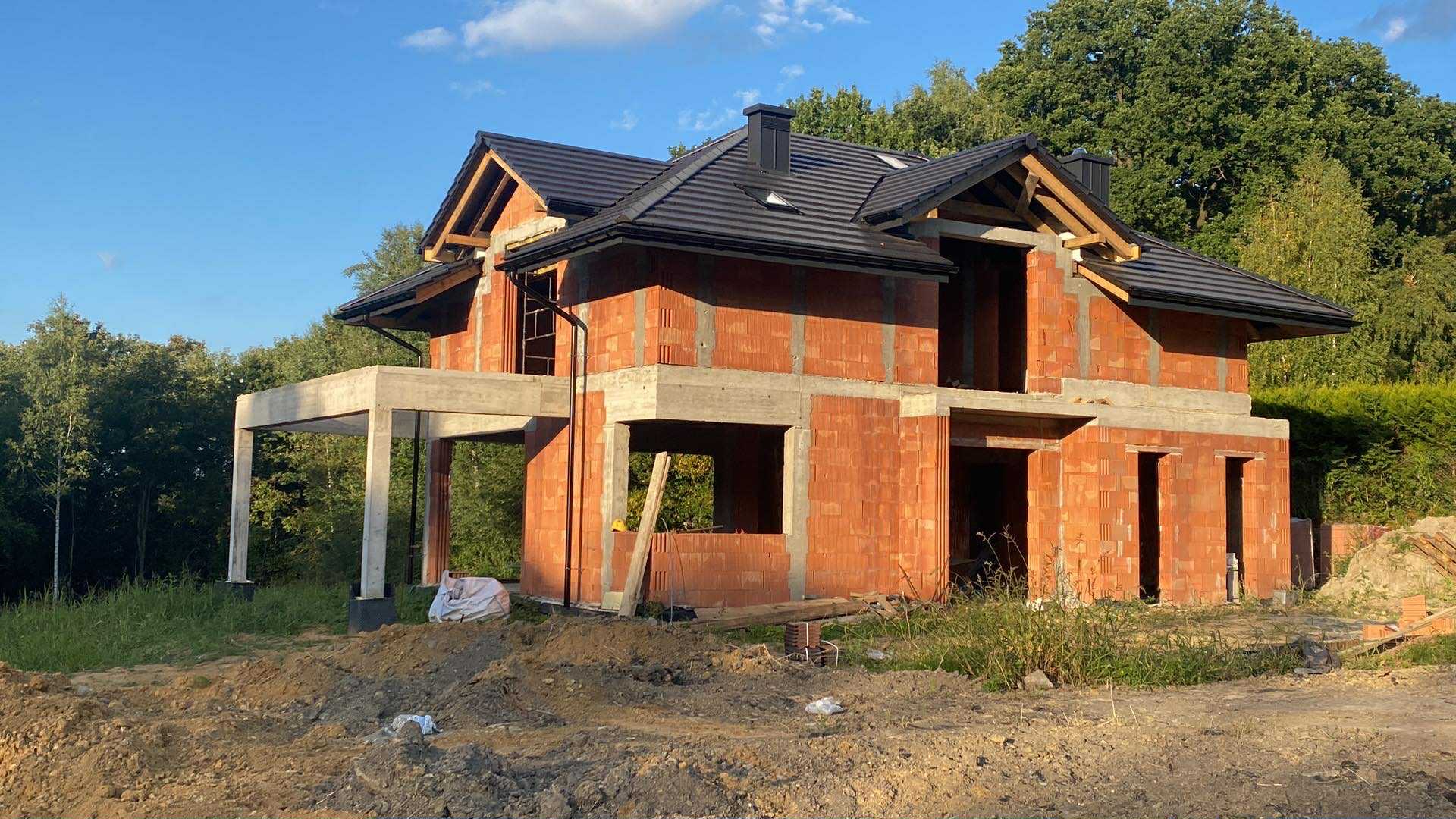 Surowy, dwukondygnacyjny dom z czerwonej cegły w trakcie budowy, z czarnym dachem i betonowym balkonem, otoczony zielenią i ziemią.