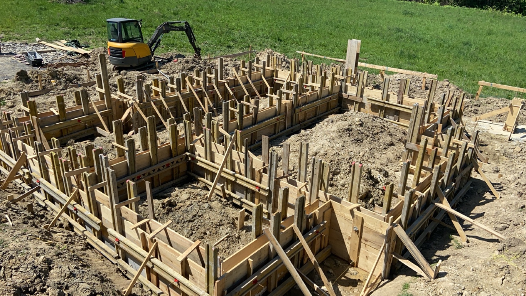 Szczegółowe ujęcie drewnianego szalunku fundamentów domu jednorodzinnego, z widoczną koparką w tle, w słoneczny dzień. Konstrukcja przygotowana do zalania betonem.