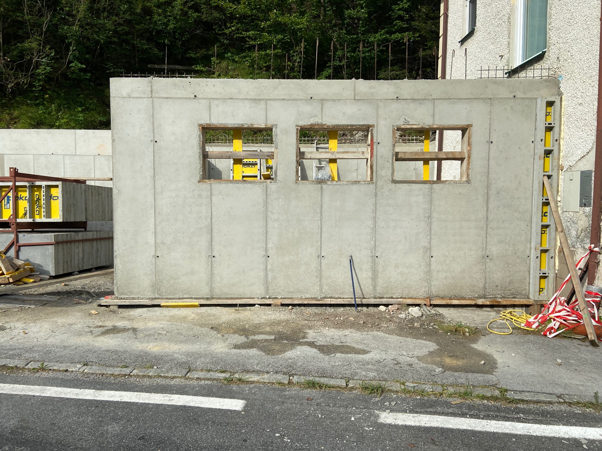 Ściana budynku żelbetowego w trakcie budowy z trzema otworami okiennymi, widoczne żółte elementy konstrukcyjne i szalunki. Na pierwszym planie fragment drogi.