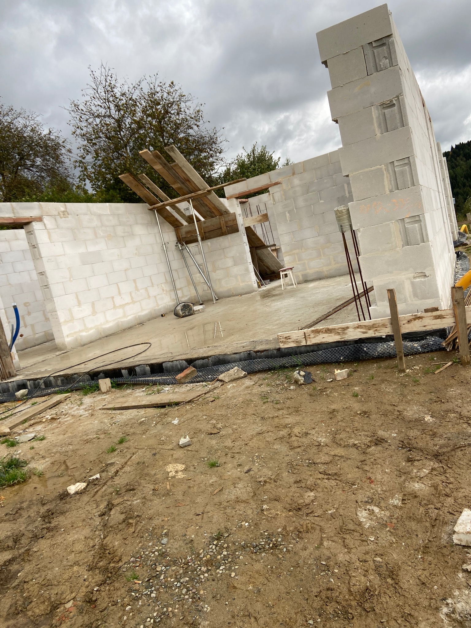 Wnętrze surowego domu w trakcie budowy z betonu komórkowego. Widoczne ściany, drewniane podpory stropu, folia izolacyjna i błotniste podłoże. Pochmurne niebo w tle.