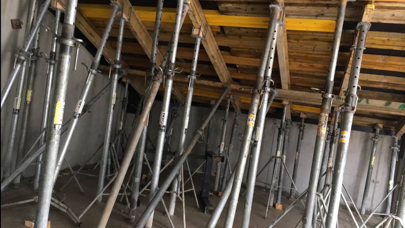 Szare, metalowe stemple budowlane podtrzymujące drewniany szalunek stropu. Widoczne żółte elementy konstrukcyjne. Ściany w tle szare, surowe.