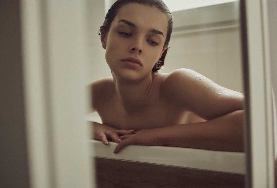 Portret kobiety z mokrymi włosami, opierającej się o brzeg wanny, uchwycony zza framugi drzwi, tworzący intymną i subtelną atmosferę.