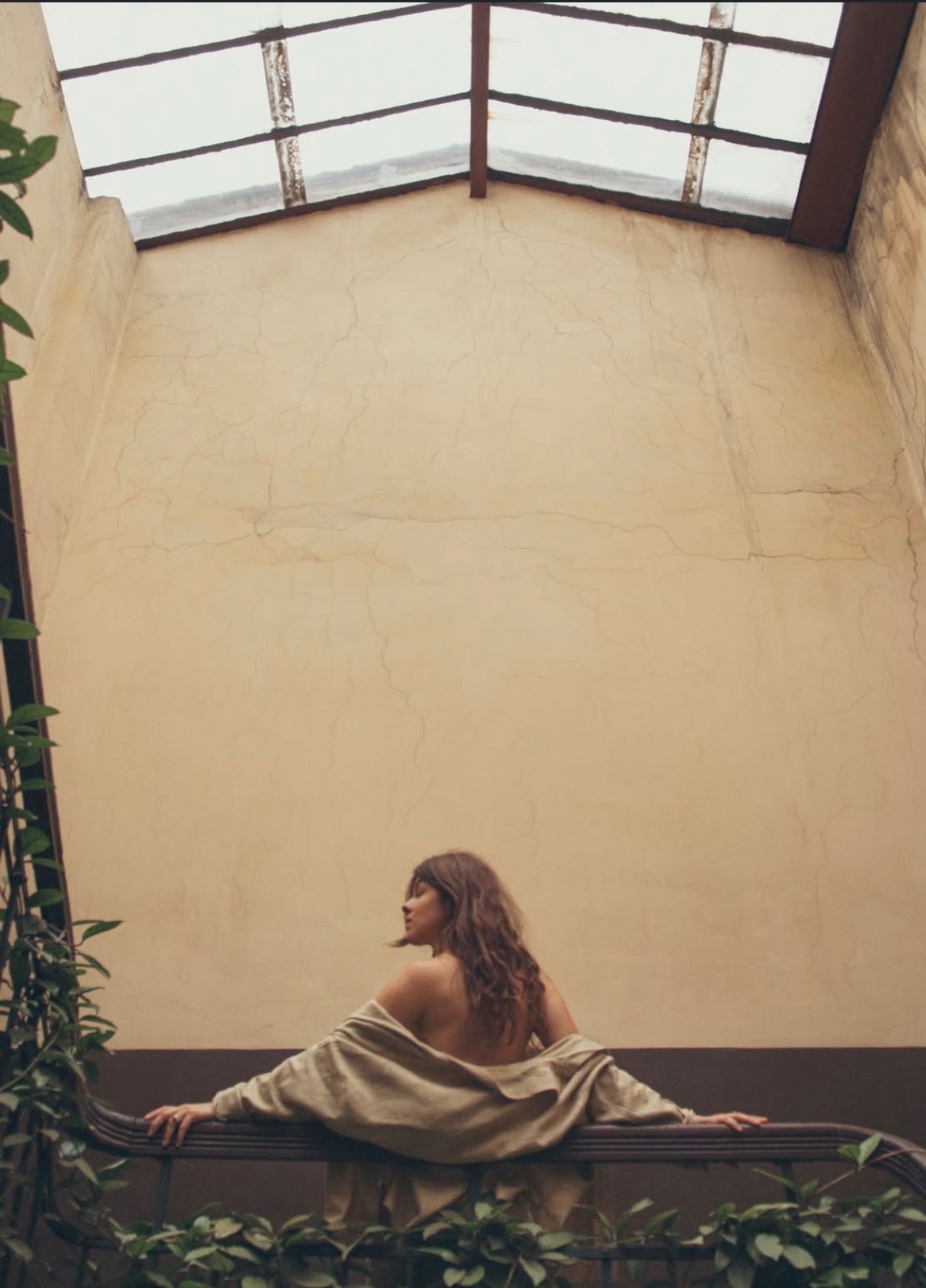 Kobieta z odkrytymi plecami opiera się o balustradę z roślinami na tle wysokiej ściany z oknem dachowym w Żywcu. Ujęcie z dołu, ciepłe kolory.