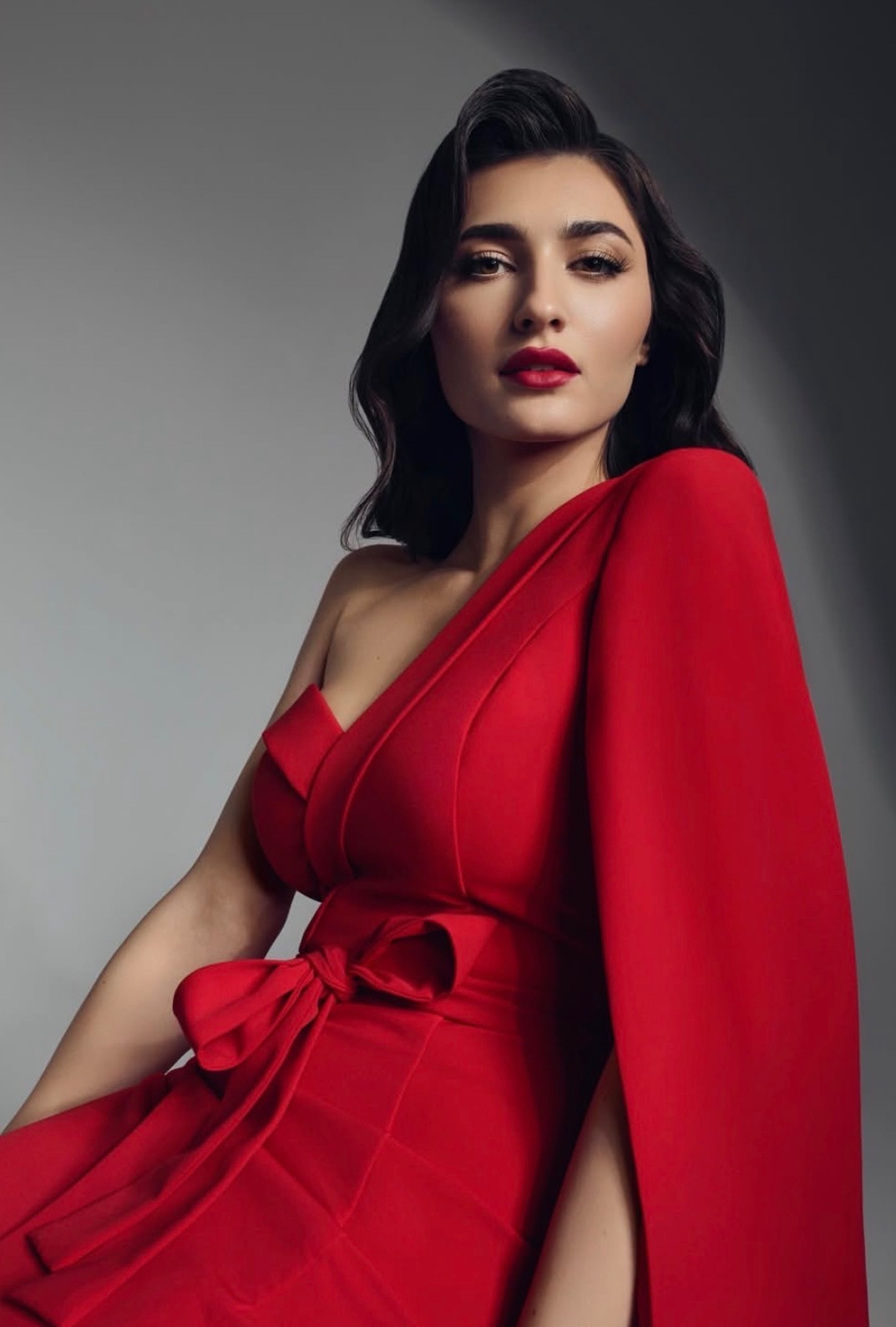 Portret kobiety w czerwonej sukni z odkrytym ramieniem, z ciemnymi włosami i czerwonymi ustami, na szarym tle. Elegancka stylizacja i profesjonalny makijaż.
