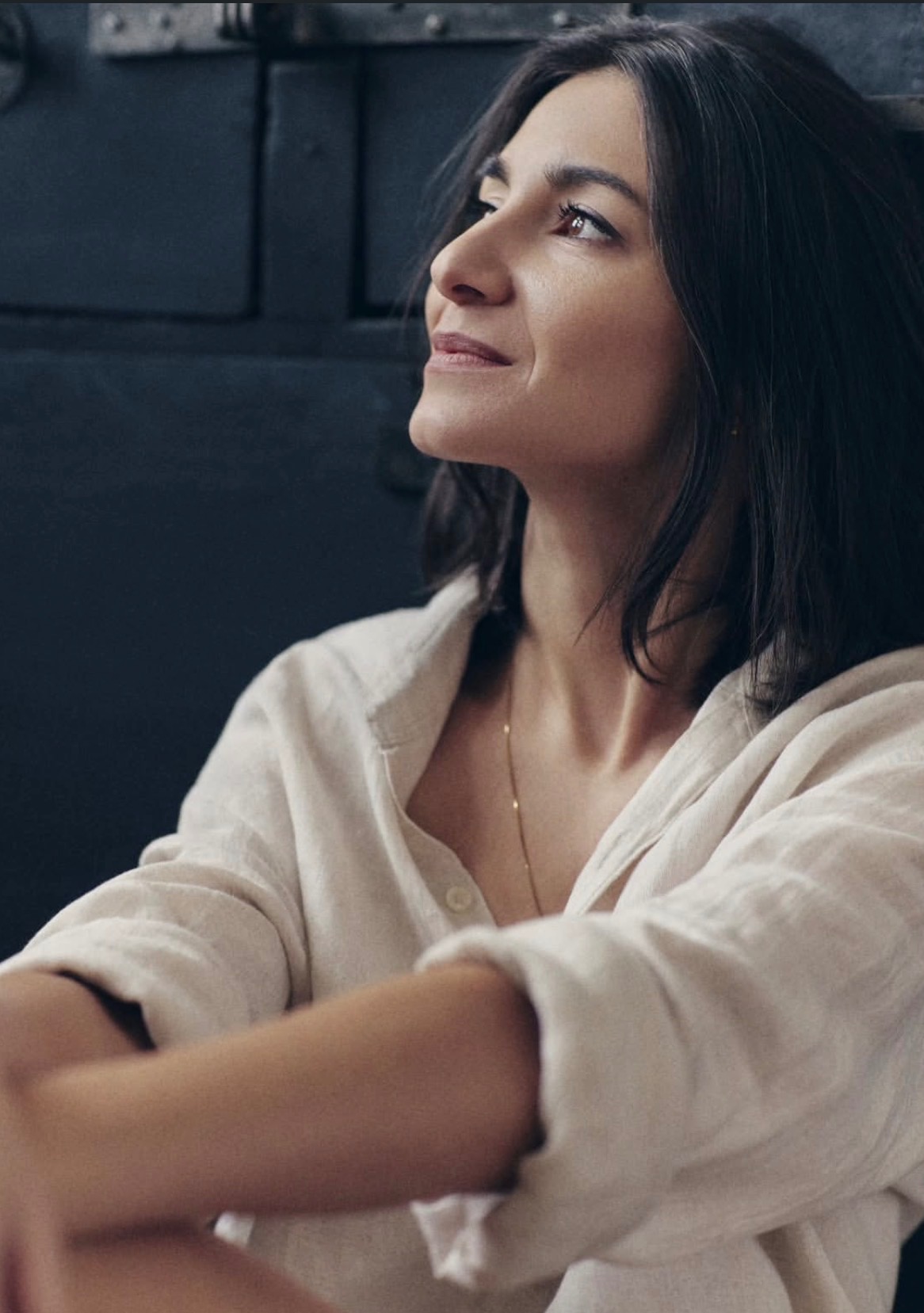 Portret kobiety z ciemnymi włosami, ubranej w lnianą koszulę, spoglądającej w górę. Delikatne światło podkreśla rysy twarzy na tle ciemnej ściany.