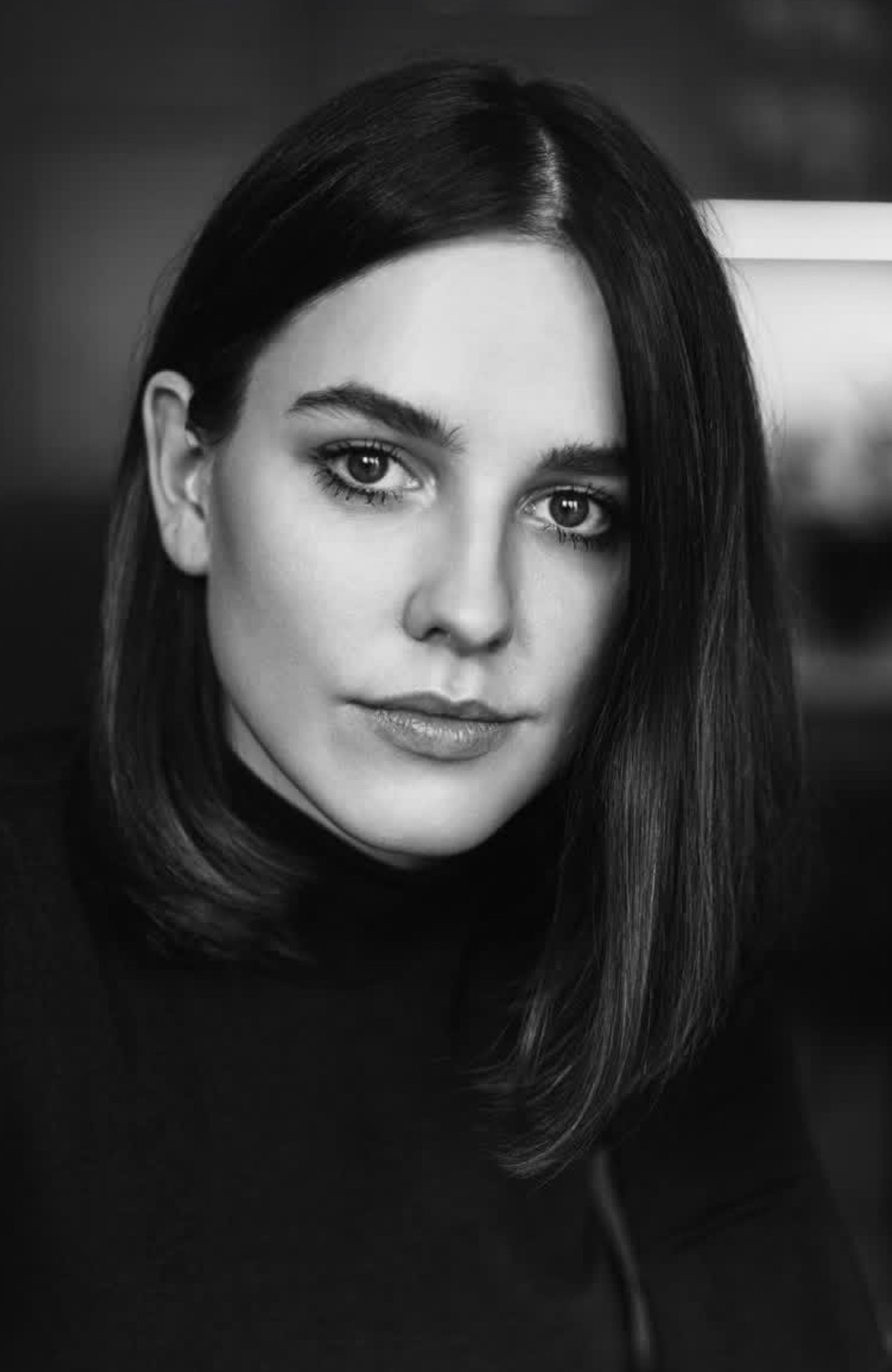 Czarno-biały portret kobiety z krótkimi, ciemnymi włosami, ubrana w czarny golf, z delikatnym makijażem i poważnym wyrazem twarzy, oświetlona miękkim światłem.