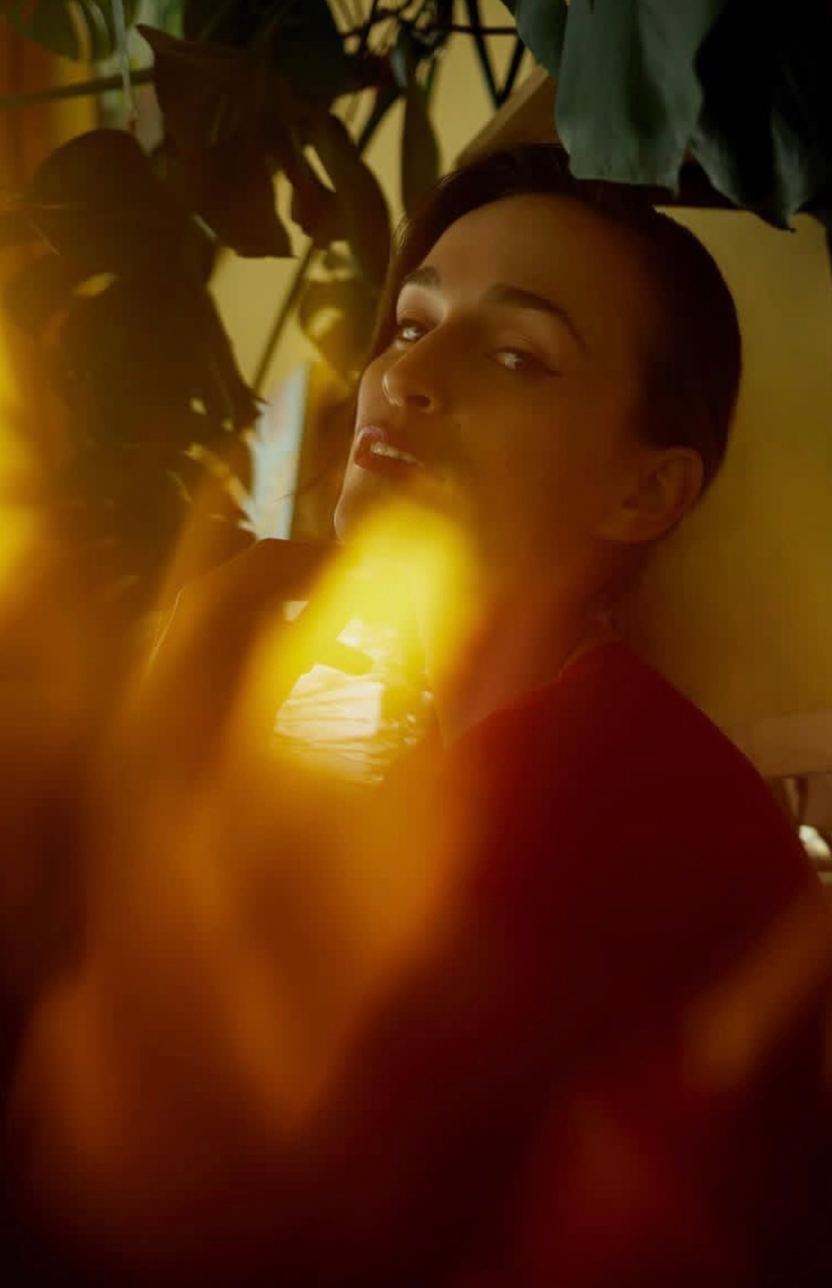 Portret kobiety w czerwonej bluzce, z roślinami w tle i efektem świetlnym, uchwycony w ciepłej tonacji, z delikatnym uśmiechem i spojrzeniem w górę.