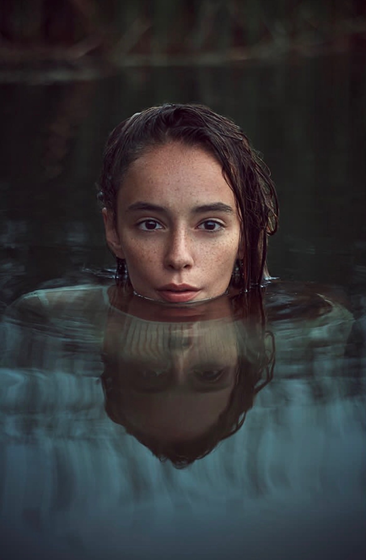 Portret kobiety zanurzonej w wodzie po szyję, z widocznym odbiciem twarzy w tafli. Delikatne światło podkreśla piegi i mokre włosy.