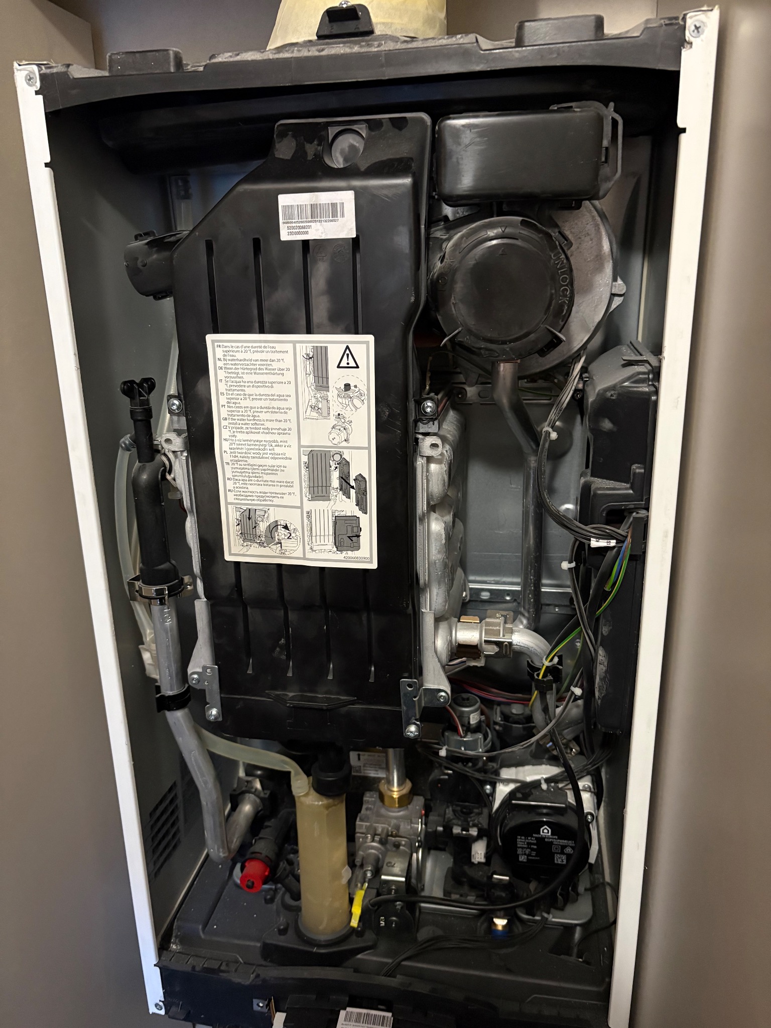 Wnętrze otwartego, białego pieca gazowego z widocznymi elementami instalacji, przewodami i naklejką informacyjną. Ujęcie z bliska, oświetlenie naturalne.