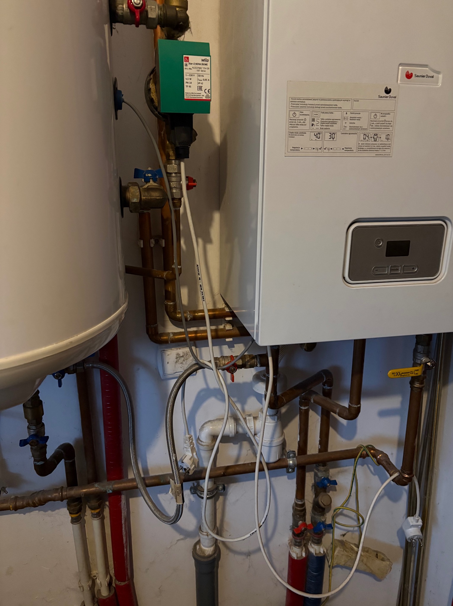 Biały piec gazowy Saunier Duval z widocznymi rurami miedzianymi, zaworami i elementami instalacji. Widoczna etykieta informacyjna i panel sterowania pieca.