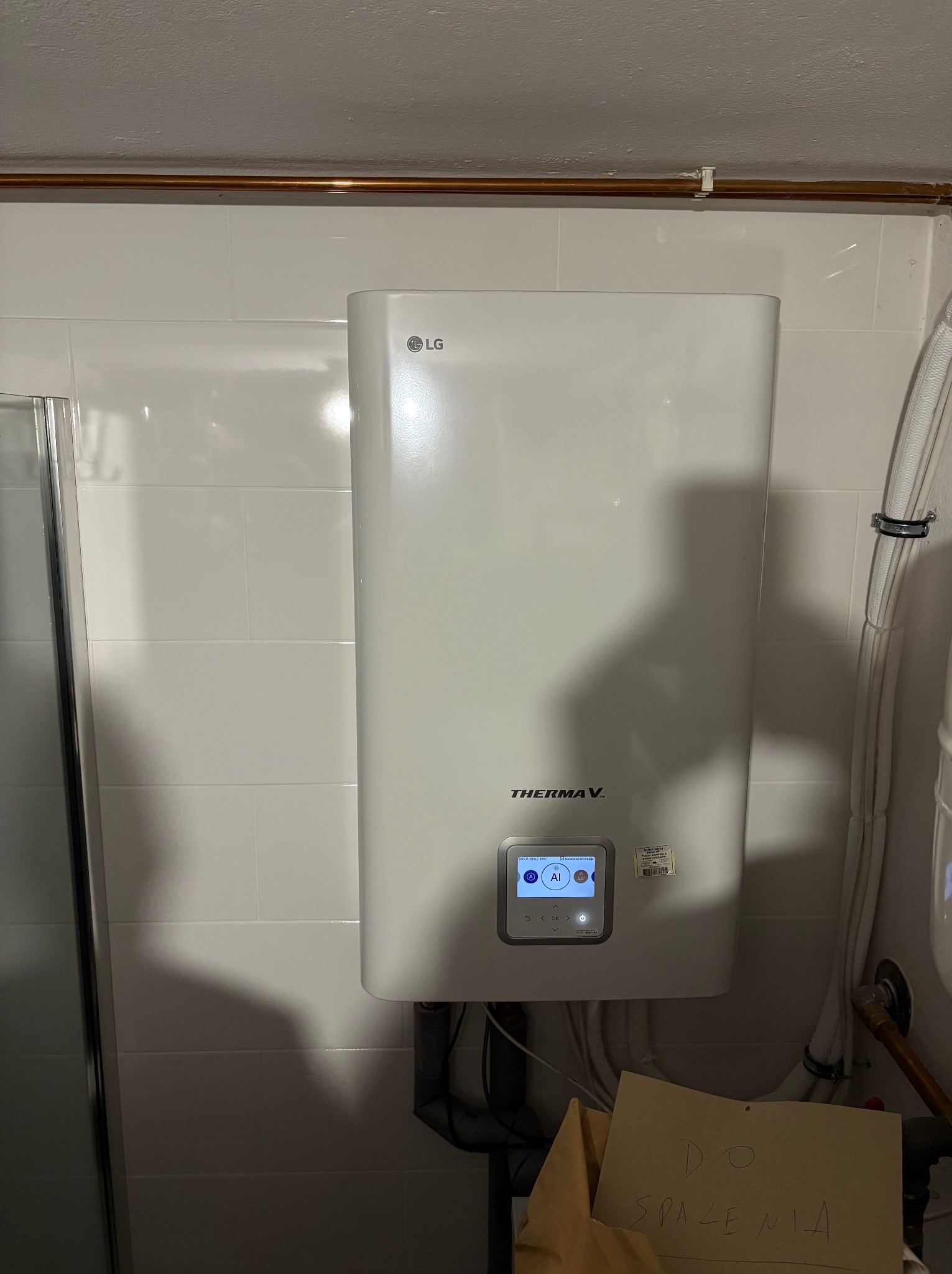 Biały, wiszący piec gazowy LG Therma V na tle białych płytek. Widoczne rury, wyświetlacz pieca i kartka z napisem 'Do Spalenia'.