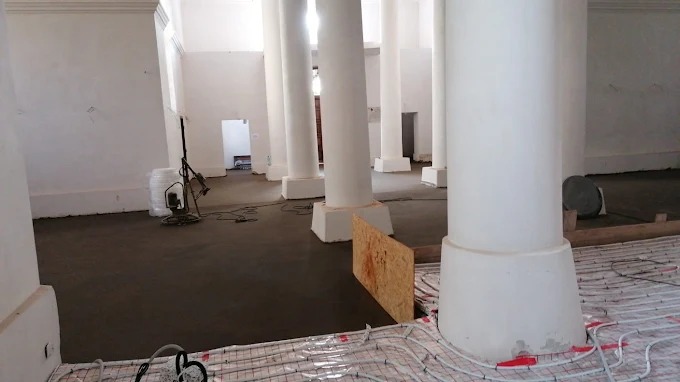 Wnętrze z kolumnami i wylaną, szarą posadzką. Widoczne ogrzewanie podłogowe, narzędzia i płyta OSB. Jasne pomieszczenie w trakcie prac wykończeniowych.