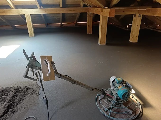 Świeżo wylana, gładka posadzka w surowym wnętrzu poddasza z drewnianą konstrukcją dachu. Na pierwszym planie maszyna do zacierania posadzek.