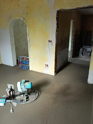 Wyrównywanie wylewki maszyną w pomieszczeniu z żółtymi ścianami. Widoczne kable wystające ze ściany i przejście do innego pokoju. W tle regał z rurami.