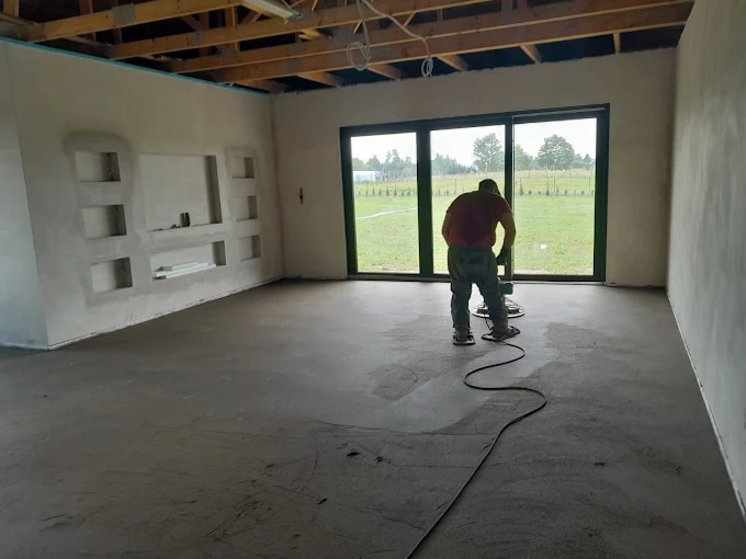 Szlifowanie wylewki w przestronnym wnętrzu z dużym oknem tarasowym. Widoczna surowa ściana z wnękami i drewniana konstrukcja stropu.