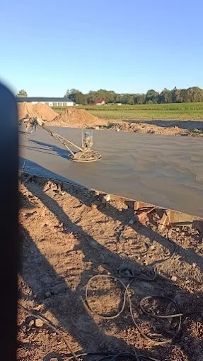 Zacieranie betonu zacieraczką mechaniczną na budowie. Widoczne przewody i ziemia. W tle budynki i zieleń. Ujęcie z dołu, widać cień.