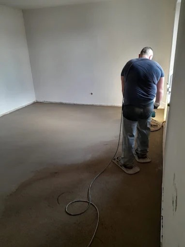 Pracownik w nakładkach na buty wyrównuje świeżo wylaną, szarą wylewkę w jasnym pomieszczeniu. Widoczny kabel od szlifierki i gładkie ściany.