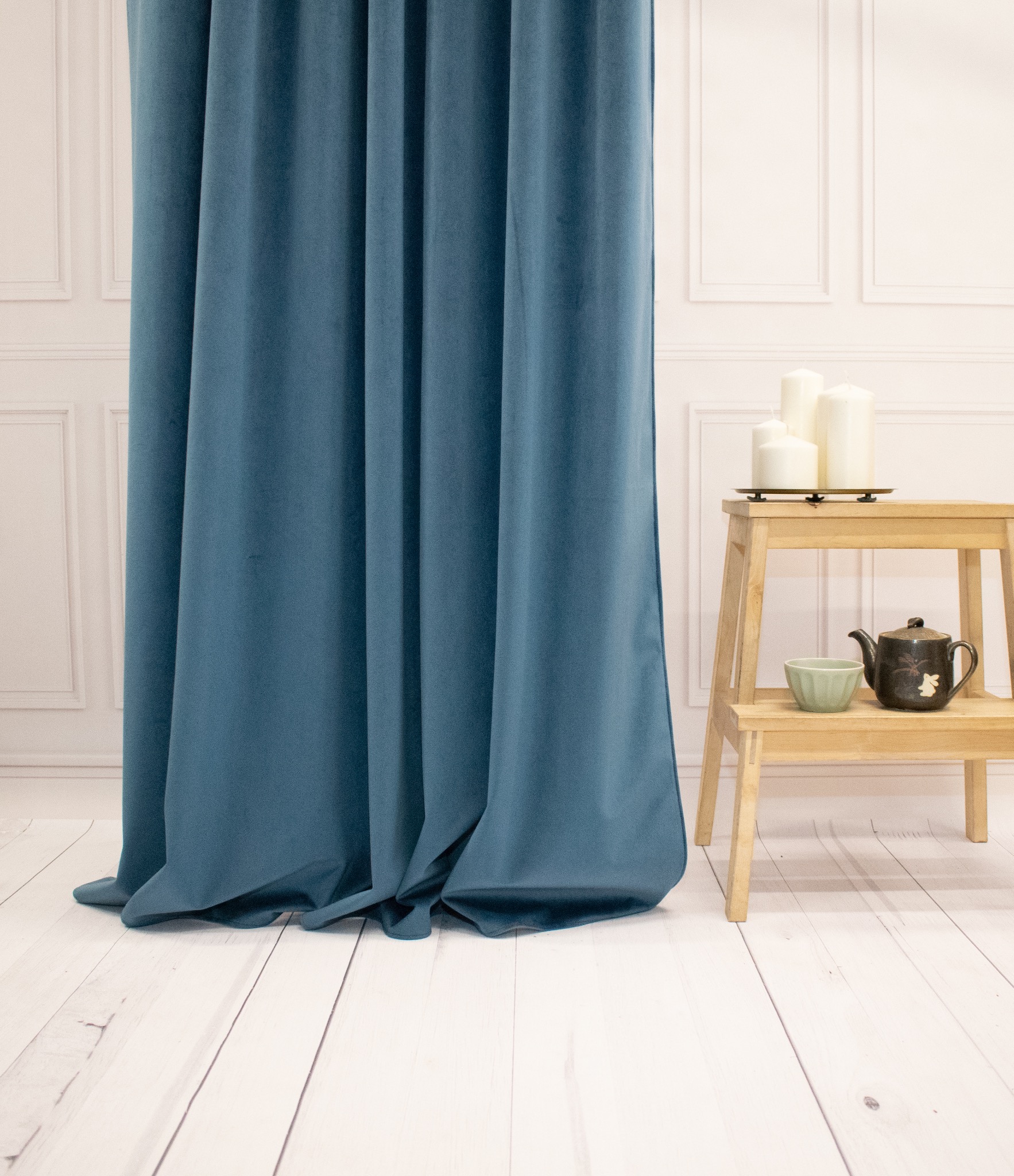 Turkusowa, welwetowa zasłona w eleganckim wnętrzu. Obok drewniany stołek z dekoracjami: świece, czajnik i miseczka. Jasna podłoga i ściana z panelami.