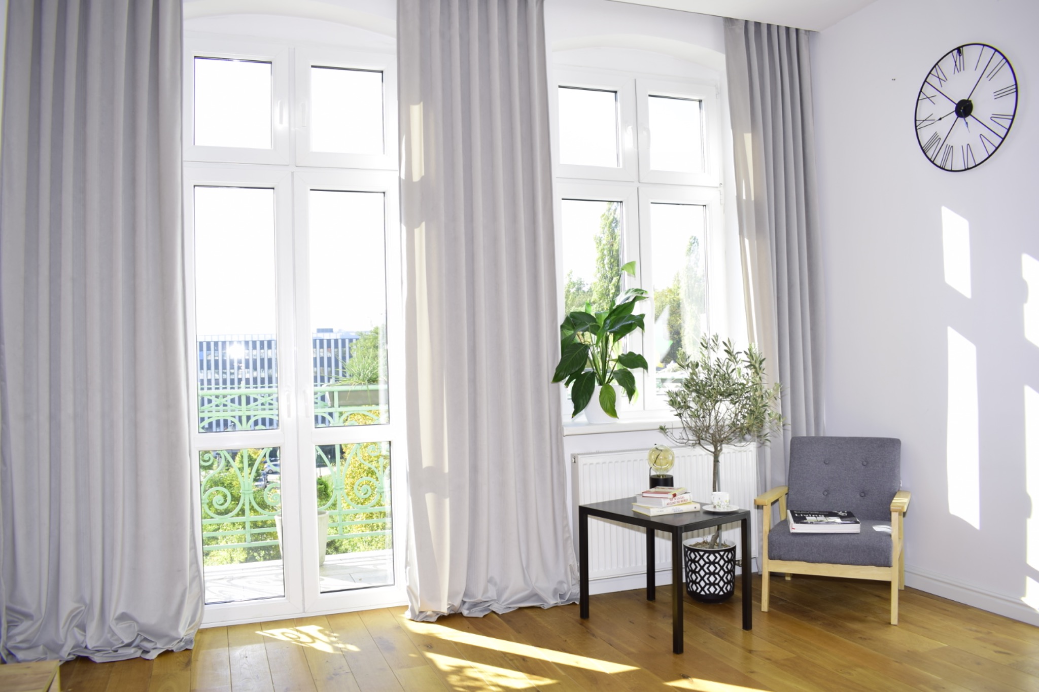 Jasne wnętrze z oknami przysłoniętymi długimi, szarymi zasłonami. Widoczny fragment balkonu z zieloną balustradą i element dekoracyjny - zegar na ścianie.
