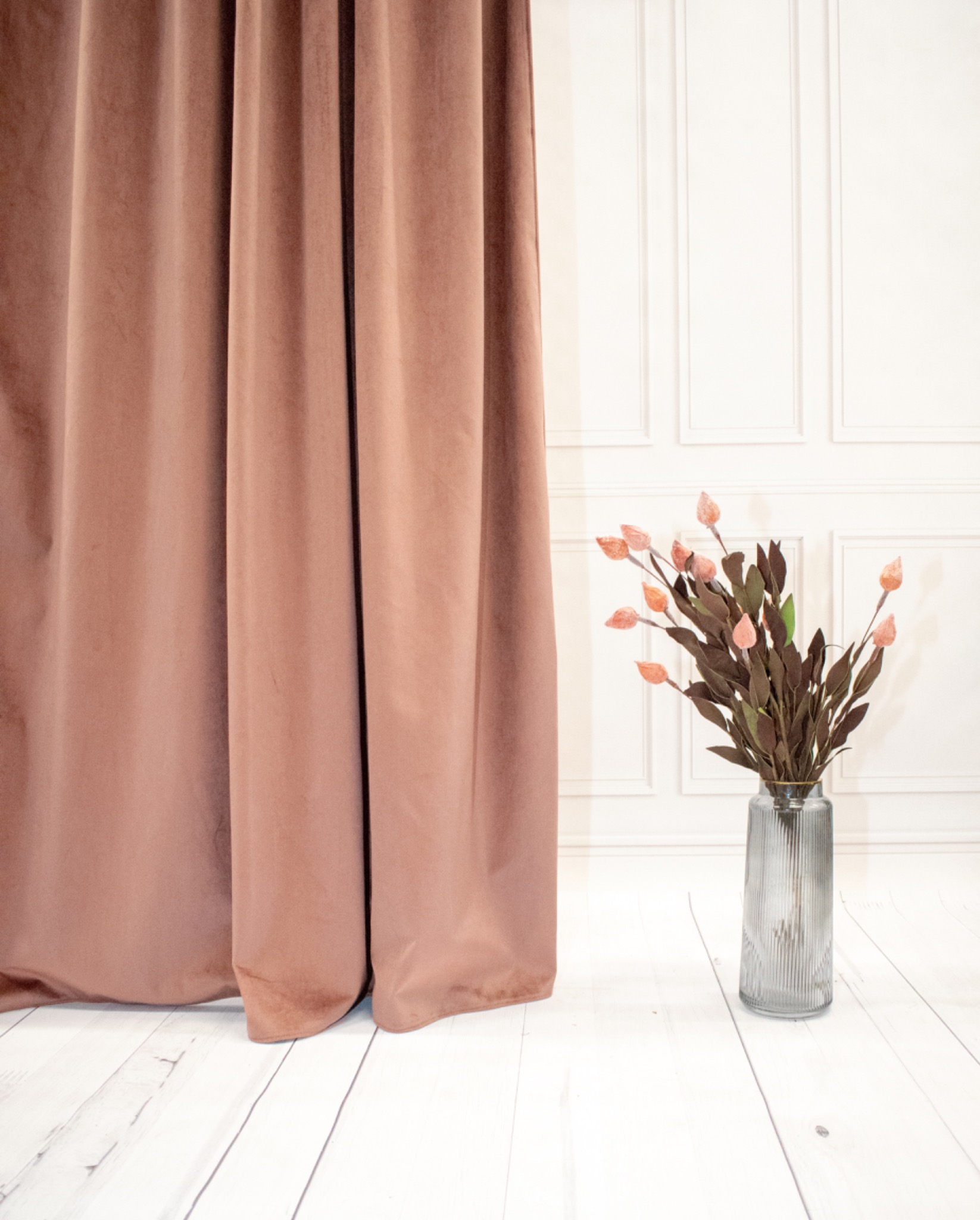 Beżowa, welurowa zasłona na tle białej ściany z dekoracyjnymi panelami i wazonem z kwiatami. Ujęcie z dołu, podkreślające fakturę materiału i elegancję wnętrza.