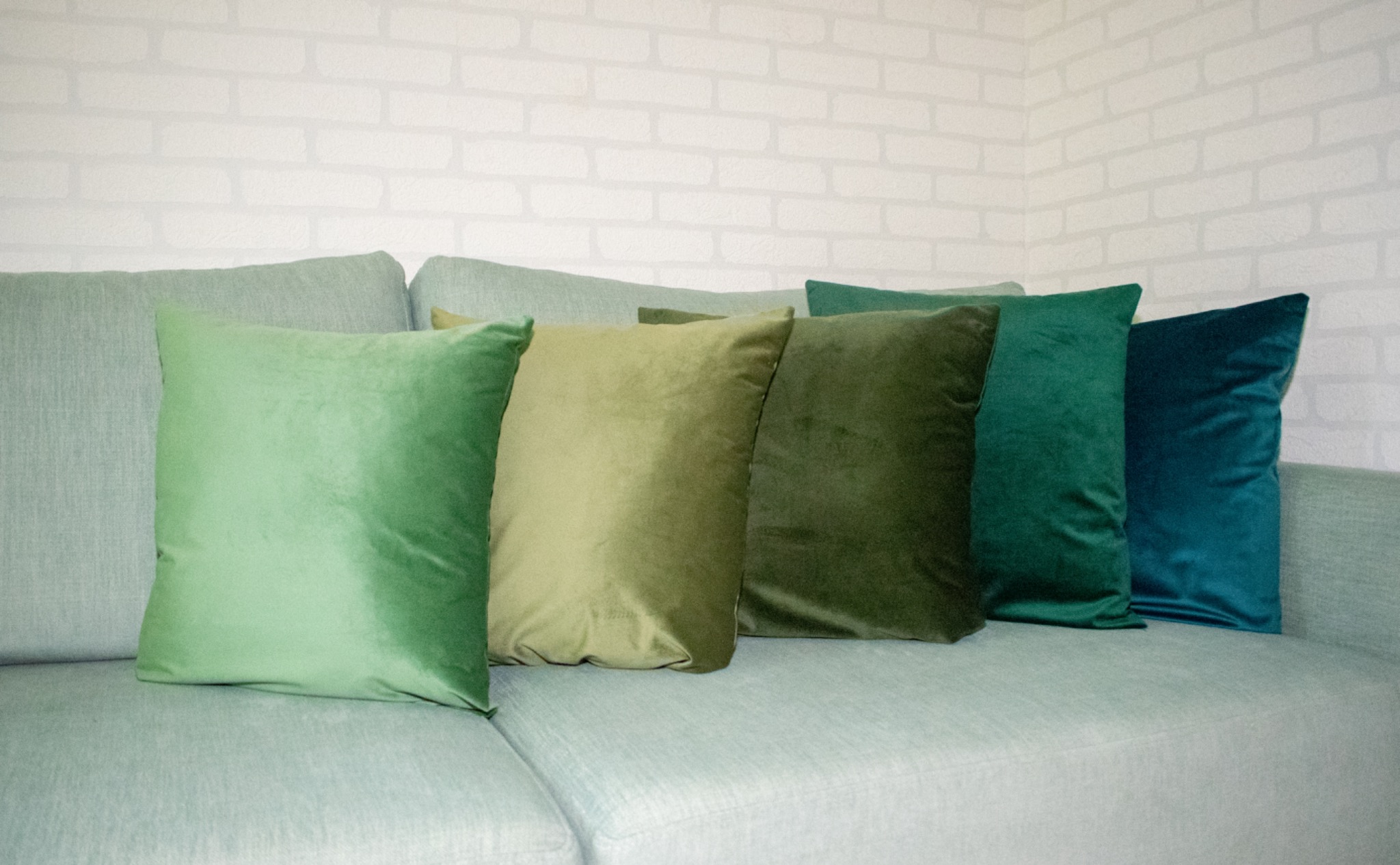 Dekoracyjne, welurowe poduszki w odcieniach zieleni i turkusu na jasnej sofie. Tło stanowi ściana z białej cegły. Stonowana kolorystyka, miękkie światło.