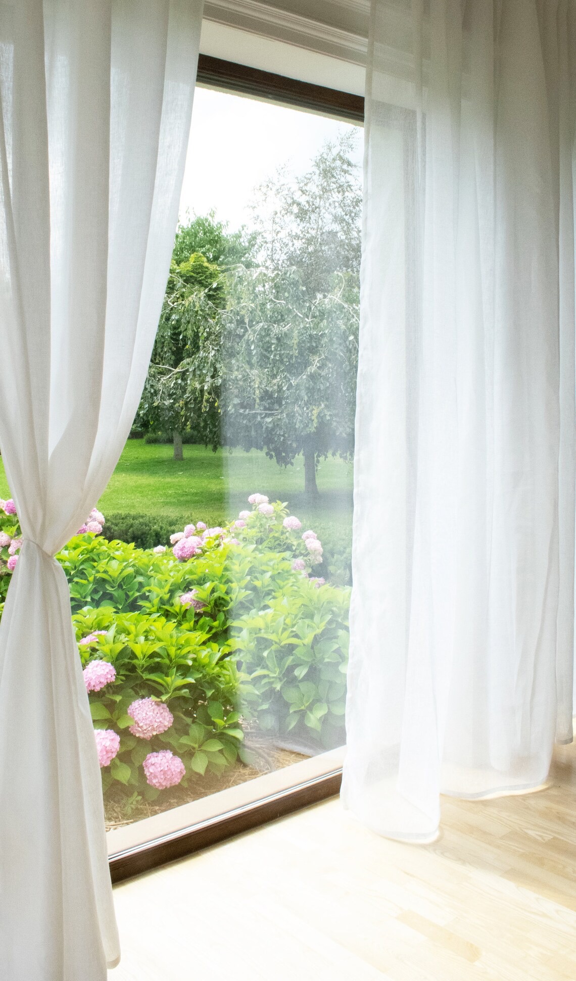 Delikatne, białe zasłony w oknie z widokiem na kwitnący ogród z różowymi hortensjami. Jasne, naturalne światło podkreśla subtelność tkaniny.