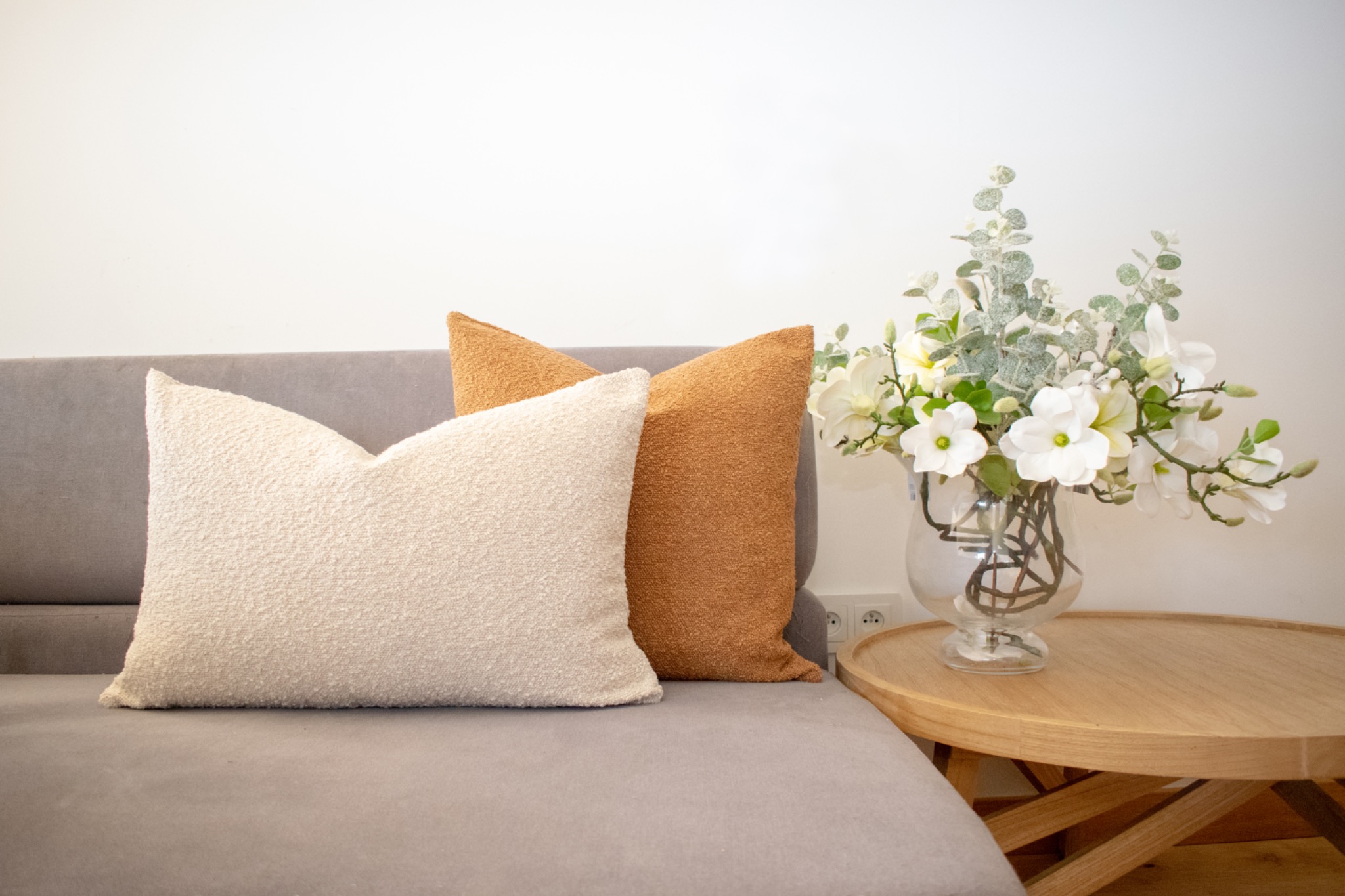 Dekoracyjne poduszki z fakturą boucle w odcieniach beżu i brązu na szarym narożniku, obok okrągły stolik z wazonem białych kwiatów. Jasne, naturalne światło.