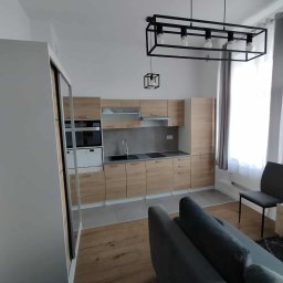 VENLOP SPÓŁKA Z OGRANICZONĄ ODPOWIEDZIALNOŚCIĄ - Jasna kuchnia z drewnianymi frontami, szarym blatem i nowoczesnym oświetleniem. Widoczny fragment sofy i szafy wnękowej. Minimalistyczny design.