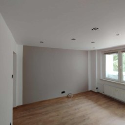 VENLOP SPÓŁKA Z OGRANICZONĄ ODPOWIEDZIALNOŚCIĄ - Jasne wnętrze po malowaniu ścian i ułożeniu paneli podłogowych. Widoczne okno z grzejnikiem, sufit z oświetleniem punktowym i fragment instalacji elektrycznej przy podłodze.