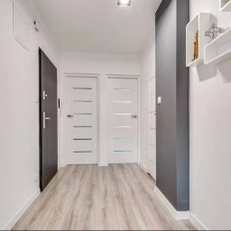 VENLOP SPÓŁKA Z OGRANICZONĄ ODPOWIEDZIALNOŚCIĄ - Nowoczesny korytarz z panelami podłogowymi, dwiema białymi drzwiami z poziomymi przeszkleniami i ciemnymi drzwiami wejściowymi. Na ścianie białe półki.