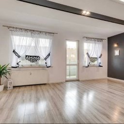 VENLOP SPÓŁKA Z OGRANICZONĄ ODPOWIEDZIALNOŚCIĄ - Jasne, przestronne wnętrze po remoncie: panele podłogowe, ceglana ściana, okna z dekoracyjnymi firanami, minimalistyczne oświetlenie sufitowe i ciemna ściana akcentująca.