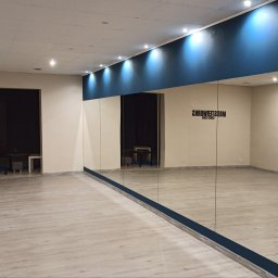 VENLOP SPÓŁKA Z OGRANICZONĄ ODPOWIEDZIALNOŚCIĄ - Wykończona sala taneczna z lustrami na ścianie, jasną podłogą imitującą drewno i niebieskim pasem na górze ściany. Widoczne oświetlenie sufitowe i logo studia na ścianie.