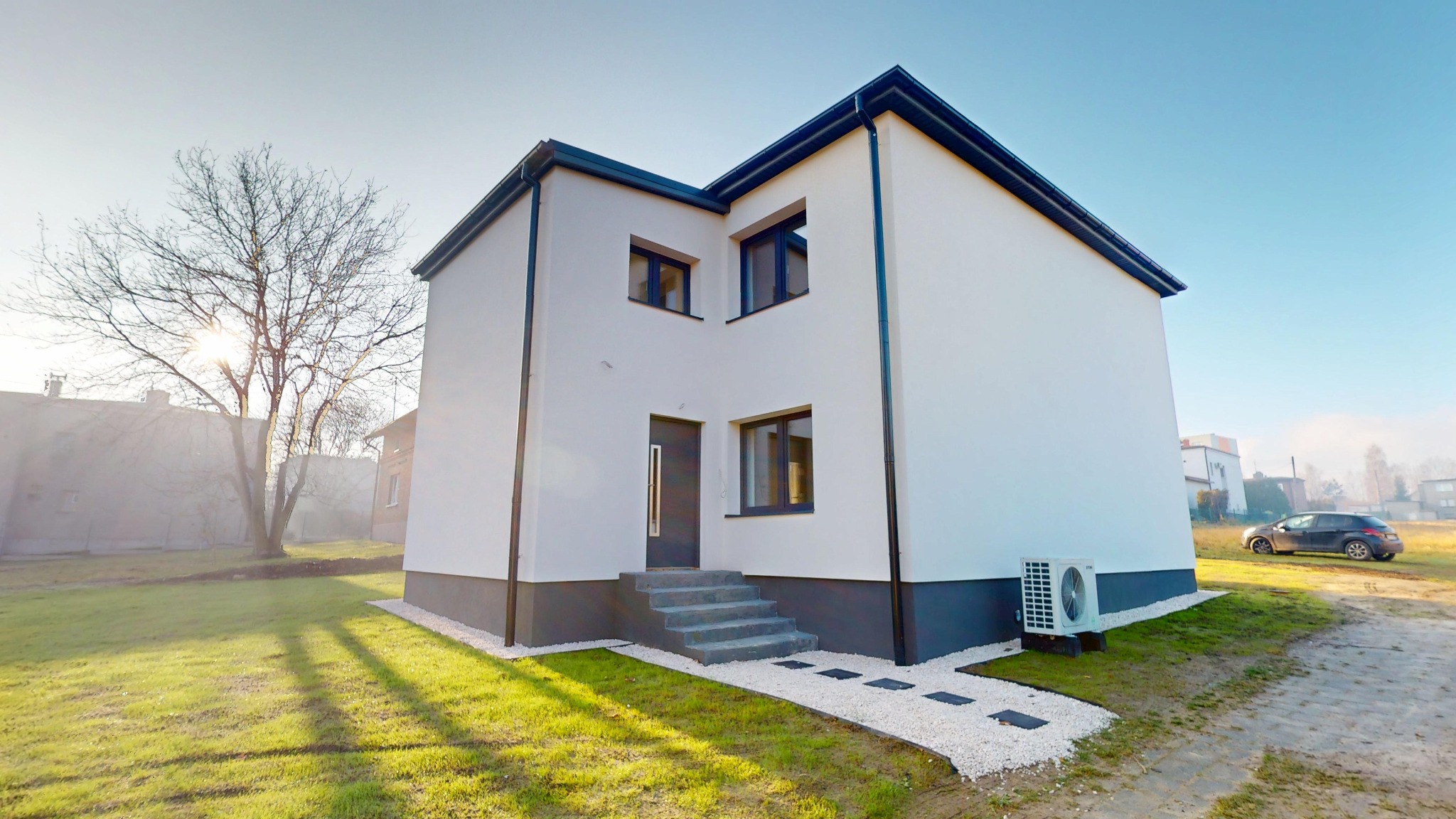 Nowoczesny, dwukondygnacyjny dom z białymi ścianami i antracytowymi detalami, widoczny z zewnątrz, z zadbanym trawnikiem i ścieżką z białego żwiru.