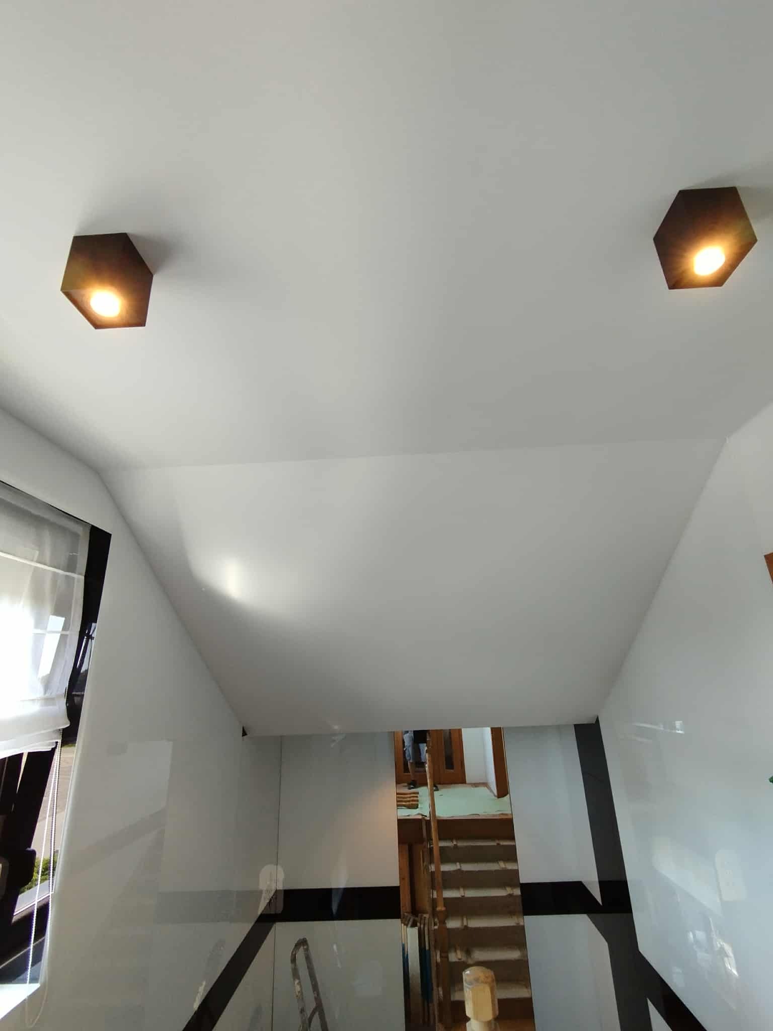 Biały sufit z dwoma sześciokątnymi lampami w ciepłym odcieniu, widok na schody i okno z żaluzjami. Minimalistyczny design wnętrza.