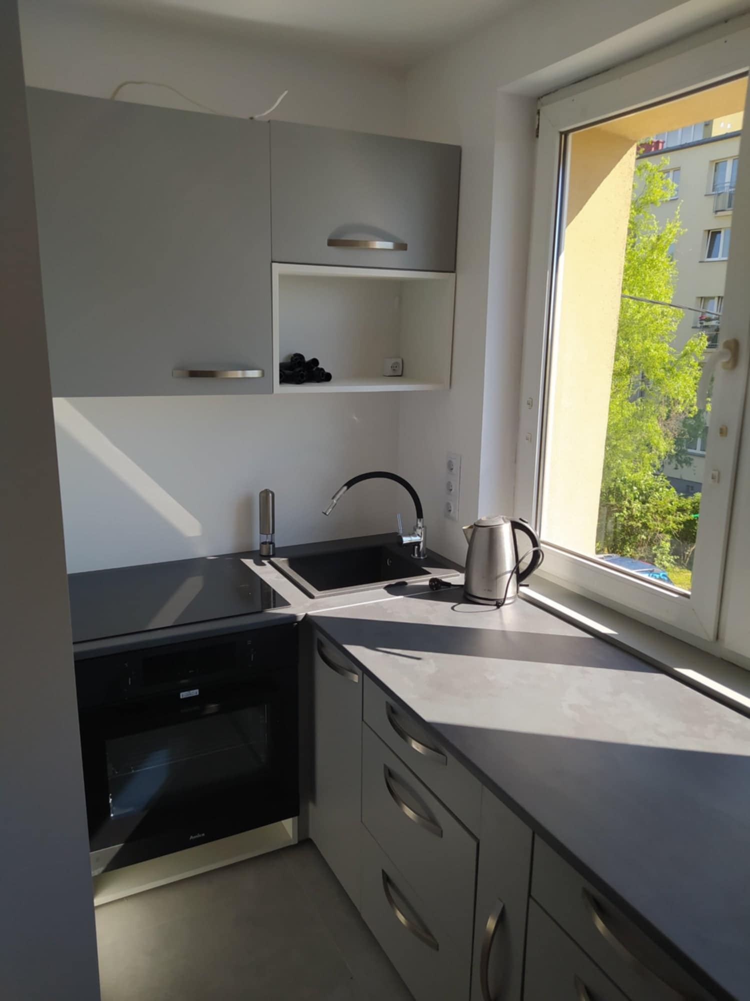 Nowoczesna, szara kuchnia z oknem, czarnym zlewem i płytą. Na blacie czajnik elektryczny. Widok na zieleń za oknem. Minimalistyczny design, jasne wnętrze.